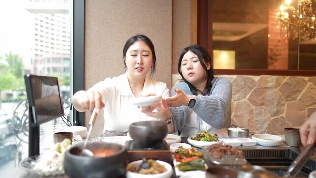 Two women enjoy Korean barbecue together. They smile, savoring delicious, steamy dishes. Warm lighting enhances cozy, intimate restaurant vibes. Great for food, culture, or lifestyle content