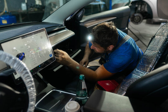 Car cabin shows large touchscreen and open glove box in workshop. Technician leans in probing wiring with flashlight testing menu controls