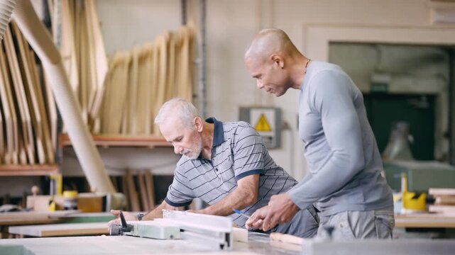 Two carpenters working together in a wood workshop. An older, skilled craftsman is teaching a younger worker how to measure wood planks