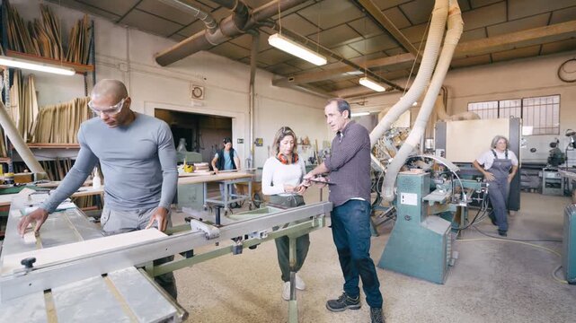 Young male apprentice learning how to use a table saw for cutting wood planks. Male and female supervisors guiding the trainee in a factory