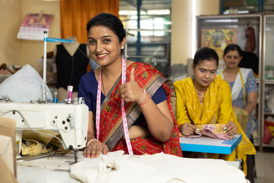 Confident skilled Indian woman tailor giving thumbs up while working at sewing machine in a textile export house. Female garment worker smiling at the camera with coworkers in the background.