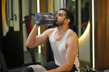 Fototapeta premium Sion, Mumbai, India-22nd March 2026-Young athletic man drinking water from a large shaker bottle after an intense workout at a modern fitness gym. Concept of hydration, post-workout recovery, healthy 