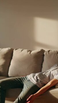 Vertical video: Entering teen in T-shirt jeans walking to sofa, reclining on pillow in sun to rest