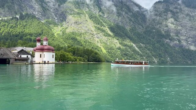 Boat, ferry arrives at St. Bartholomew stop. Panoramic, scenic view of Konigssee Lake. St. Bartholomew church and wooden boathouse. People are walking around. Konigssee, Bavaria, Germany 