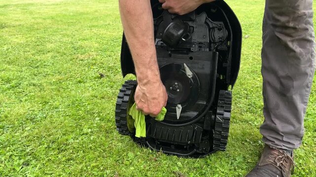 Cleaning the underside of a robotic lawn mower with a cloth after rinsing, removing stuck grass and dirt from the cutting deck as part of routine maintenance to improve cutting performance and extend 