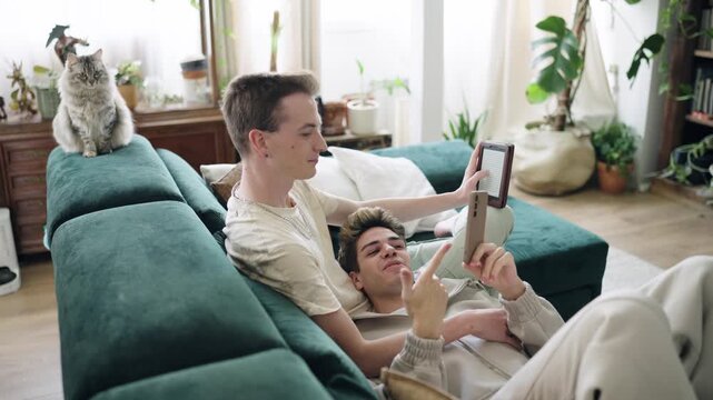 Young gay couple relaxing on the couch in their living room, using a smartphone and an e reader, with their cat sitting nearby