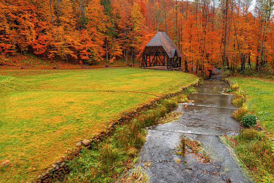 A wooden gazebo stands near a flowing stream in a park surrounded by vibrant autumn trees with orange and red foliage.