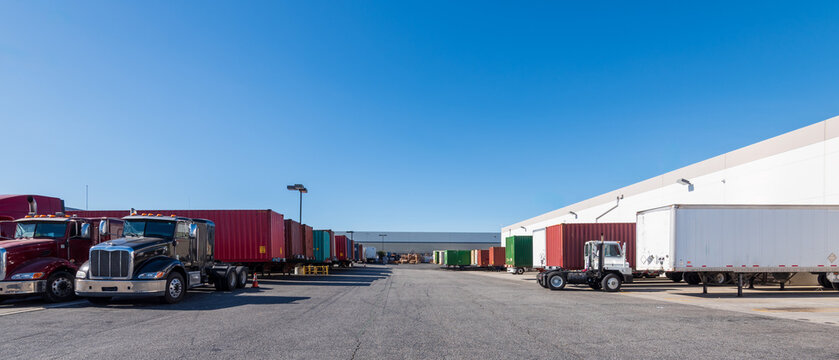 Commercial semi-trucks and shipping container trailers parked outside a large modern logistics distribution warehouse under a clear blue sky.