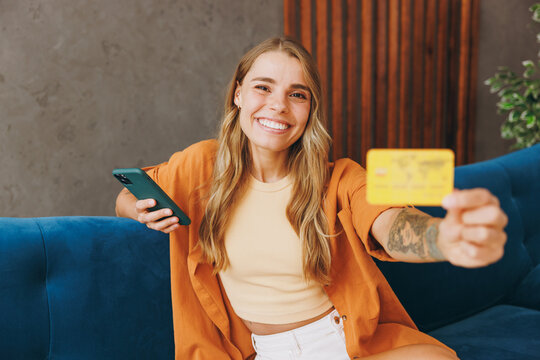 Smiling young woman wears brown shirt using mobile cell phone credit bank card shopping online order delivery sit on blue sofa stay at home hotel flat rest relax spend free time in living room indoor.