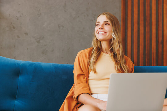 Young IT woman wears brown shirt hold use work on laptop pc computer look aside sit on blue sofa stay at home flat relax spend free spare time in living room indoors grey wall. People lounge concept.