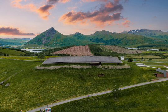 Aerial view of Lofotr Viking Museum in Borg, Lofoten, Norway, with a turf roof longhouse on a grassy ridge, excavated outlines, road, fields, peaks, at golden hour.