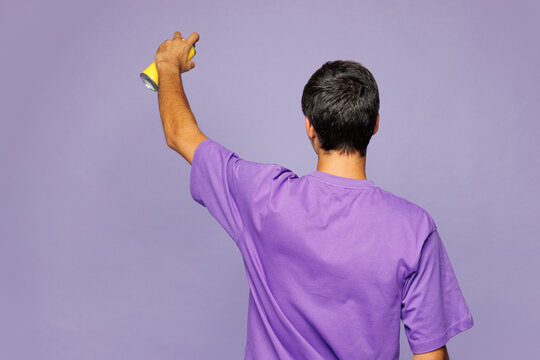 Back rear view young man he wear violet t-shirt casual clothes hold in hand spray paint bottle do graffiti stretch arm on empty wall isolated on plain pastel light purple background Lifestyle concept.