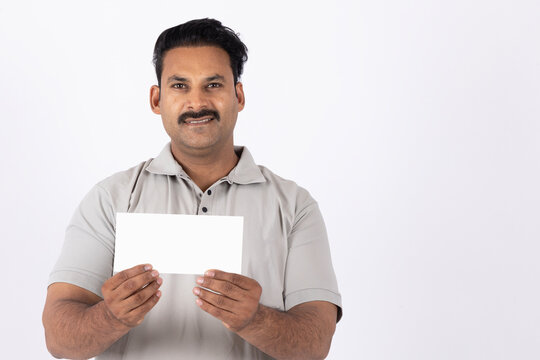 Portrait of young indian man holding bank cheque wearing t-shirt, gesturing and looking at the camera while standing against white background