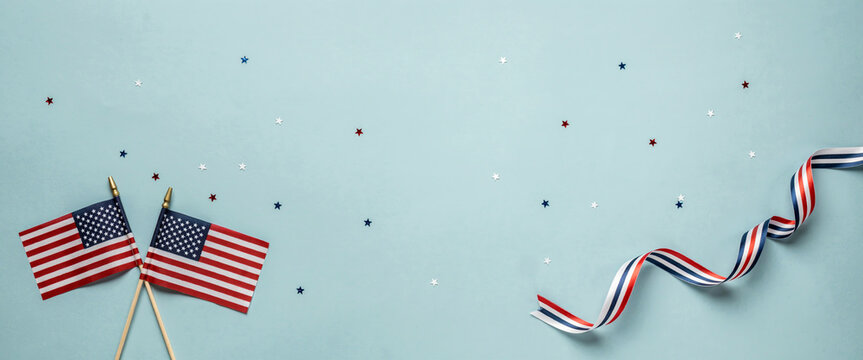 A flat lay composition featuring two small American flags crossed together with a swirling red, white, and blue ribbon against a soft light blue background.