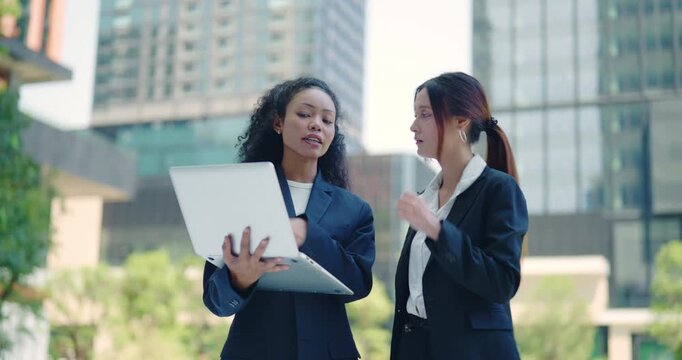 Two Businesswomen Collaborating on Laptop Outdoors in Modern Urban Business District, Diverse Professional Team Discussing Strategy Together