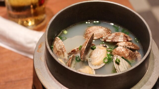 close up of Sake Steamed Clams in a pot served in a traditional metal pot, featuring fresh clams with open shells in a clear, savory broth, garnished with chopped green onions with copy space
