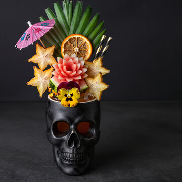 tropical cocktail in skull-shaped glass with exotic fruits and umbrella against dark background