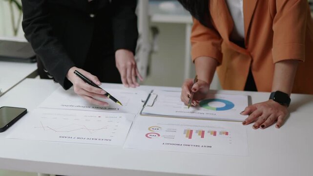 Top view of two businesswomen analyzing printed reports and charts on a desk, discussing sales and performance data together during a brainstorming meeting.