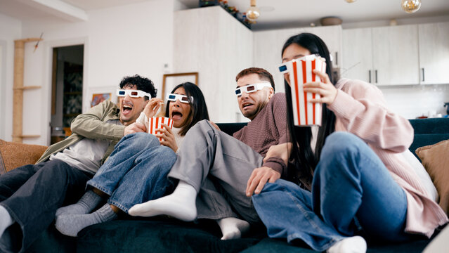 Friends sitting on a couch wearing 3D glasses, eating popcorn, and reacting to a scary film during a home cinema movie night