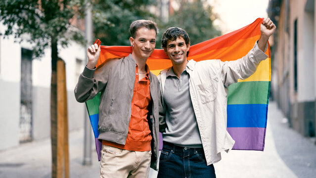 Two smiling men embracing, holding a LGBT rainbow flag outdoors, symbolizing love, equality, and pride