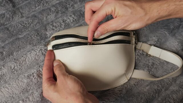 Male hands close the black zipper on a stylish white leather fanny pack resting on a grey plush surface. Concept of travel security, fashion accessories, and lifestyle organization.