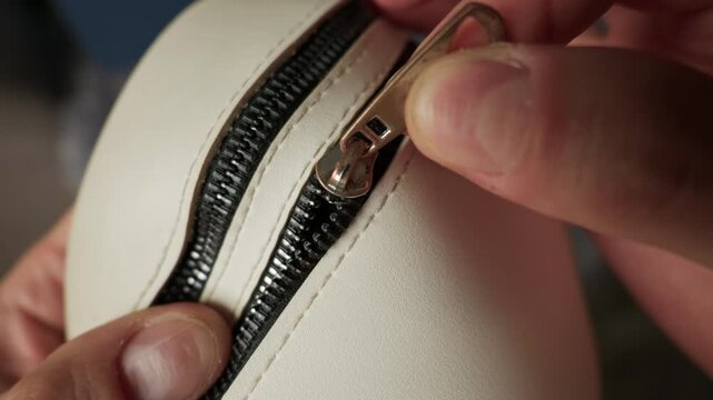 Hands firmly pull a small metal zipper slider in an attempt to close a white leather cosmetic bag. Concept of daily routines, securing personal belongings, and fixing broken accessories.
