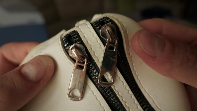 Two metallic zippers on a cream leather handbag are shown in a detailed close-up view while being held. Concept of fashion industry, garment hardware, security, and leather accessories.