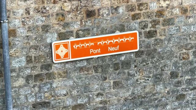 Close-up of an orange official heritage sign for "Pont Neuf" bridge mounted on a historic stone masonry wall along the Seine in Paris, France