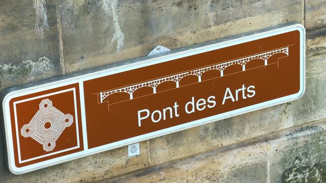 Brown metallic tourist directional sign for the "Pont des Arts" fixed onto the ancient limestone wall by the Seine River in Paris, France