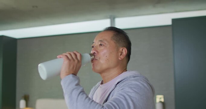 Senior Asian man feeling thirsty raising white thermos unscrewing cap drinking smiling in bedroom