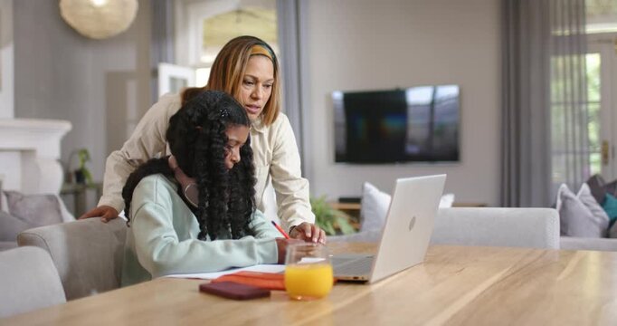 African American mom leaning over child at table pointing to notes laptop helping while kid writing