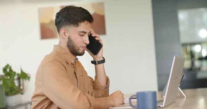 Man answering ringing phone at desk, writing on notepad for work while checking laptop