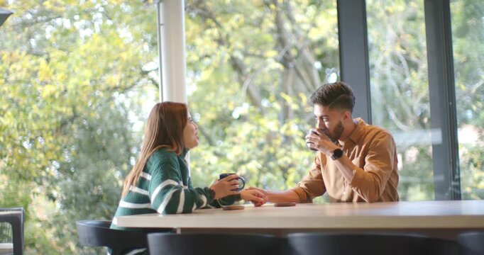 Couple sitting at table by window, woman leaning in speaking, man sipping mug touching hand sharing