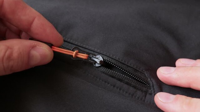 Male hands pull a bright orange zipper to close a black jacket. The person fastens the hardware preparing for outdoors. The extreme close up reveals the fine fabric texture.