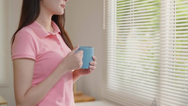 Young woman holding a blue coffee mug at home, enjoying a quiet moment by the window in a bright and cozy interior.