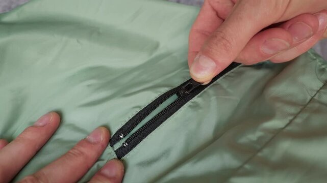 A close-up view shows a person hand carefully manipulating a small black zipper slider on a piece of light green textile fabric. Concept of garment repair, fixing broken items, and DIY.