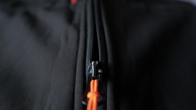 A macro view shows a black plastic zipper slider with a vibrant orange cord pull tab on dark fabric. Concept of outdoor gear, fastening hardware, manufacturing details, and color contrast.