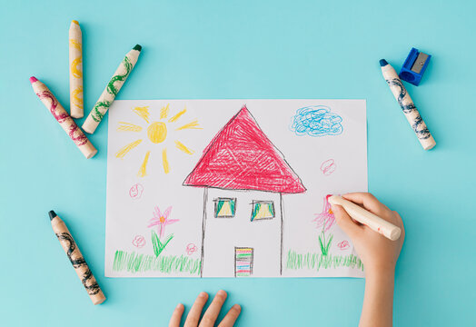 Child drawing a colorful house with crayons on paper over a blue background. Creative learning, early education, imagination, childhood art, preschool activity and back to school concept. Flat lay.