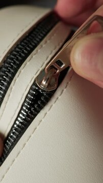 A close-up view shows a hand gripping a metal zipper pull and sliding it along black teeth on a white bag. The textured faux leather surface features clean stitching details under soft light