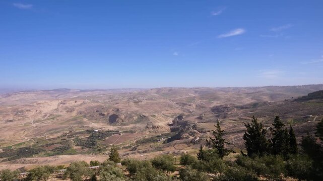 A wide panoramic view of the arid desert landscape and rolling hills as seen from the Mount Nebo lookout point in Madaba, Jordan under a clear blue sky.