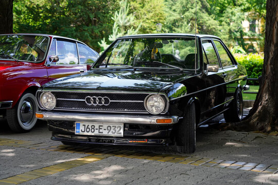 Front view of a vintage black Audi 80 B1 (Typ 80) classic two-door sedan with chrome bumper and custom front spoiler lip, parked under trees in Subotica, Serbia, on June 7, 2026