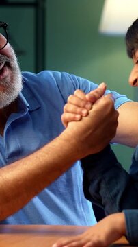 Indian grandfather and kid arm wrestling at home, both sitting together at table in modern house, elderly man and grandson enjoying playful activity, family bonding moment of two people having fun
