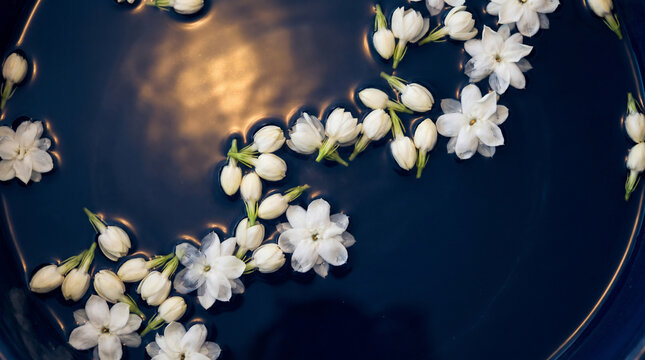 Overhead Close-up of White Thai Jasmine Flowers Floating in Dark Water Bowl with Warm Golden Candlelight Reflection