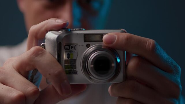 Amateur photographer holding vintage compact digital camera, pressing shutter button and taking picture. Red autofocus assist light illuminates before lens extends, flash fires, close-up, slow motion