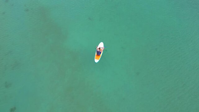 High angle minimalist drone shot of a father and his little daughter paddling together on a stand up paddleboard in vast clear turquoise sea water on a sunny day. High quality 4k footage
