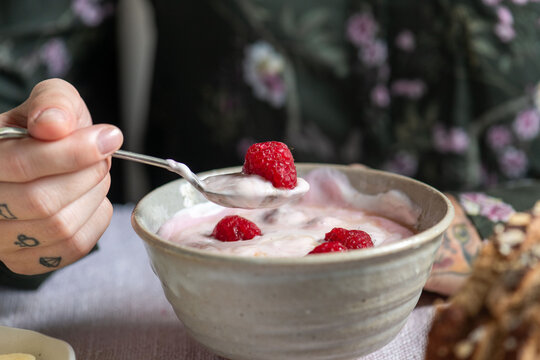 Person with tattoos enjoys yogurt with raspberries at a table.