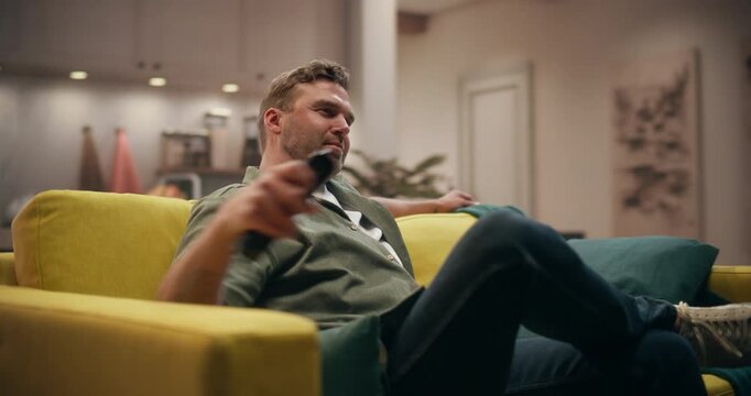 361 Degree. Man Sitting down on Couch at Home, Watching Movie on a Smart TV, Changing Channels, Enjoying Streaming Service Content and Entertainment in His Living Room. Cinematic Arc Tracking Shot.