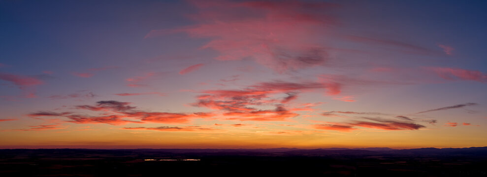 Panoramic drone view of a colorful sunset sky with pink clouds, warm orange glow and a dark horizon, ideal as a natural background or banner image. Colors: pink, orange, blue.