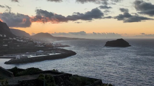 Beautiful golden sunset over the coastal town, modern harbor marina, and volcanic islet Roque de Garachico in Tenerife, Canary Islands, with dramatic evening clouds over the Atlantic Ocean.