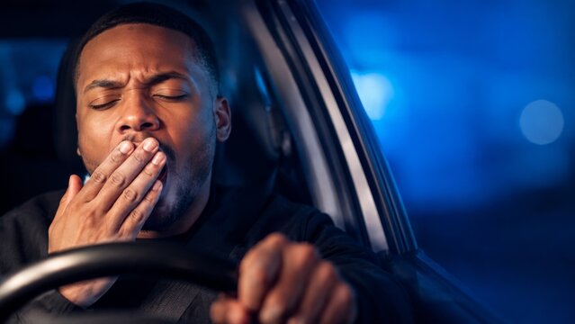 Young man yawning behind the wheel at night, showing exhaustion and fatigue while driving, highlighting the dangers of drowsy driving